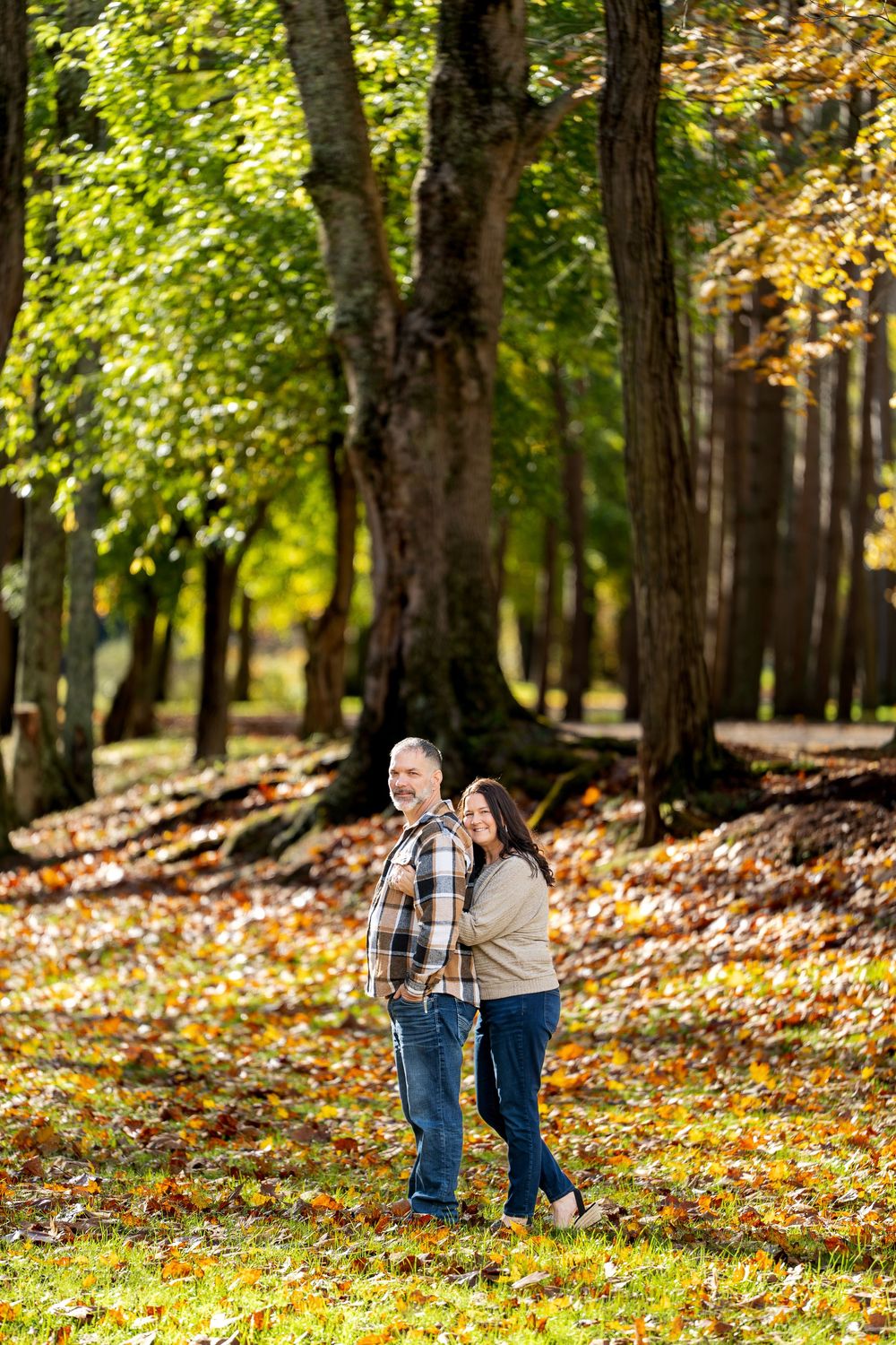 Smiling couple embracing in a sunlit autumn park surrounded by tall trees and colorful fallen leaves.
