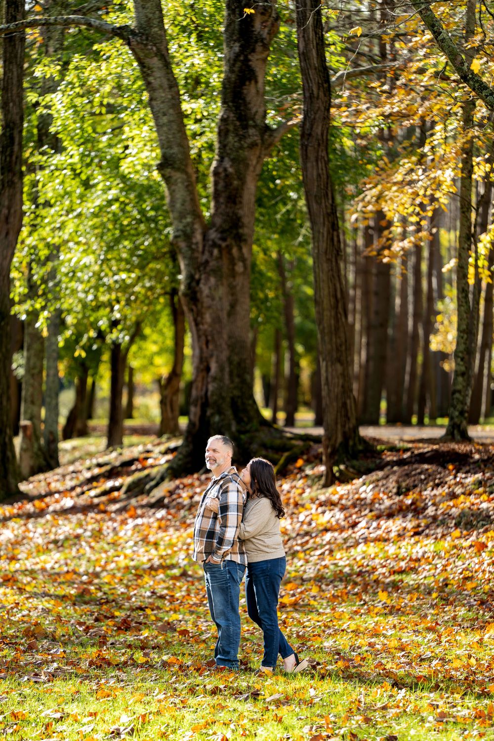 Couple in casual fall clothing stands close together on a leaf-covered lawn beneath tall trees with golden autumn foliage.