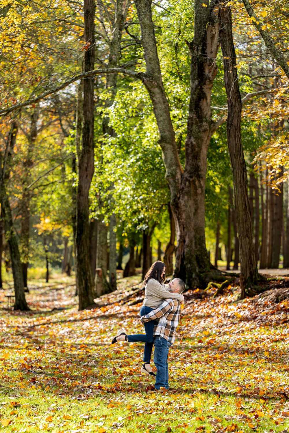 Couple twirling in a happy embrace as the man lifts his partner among tall trees and golden autumn leaves.