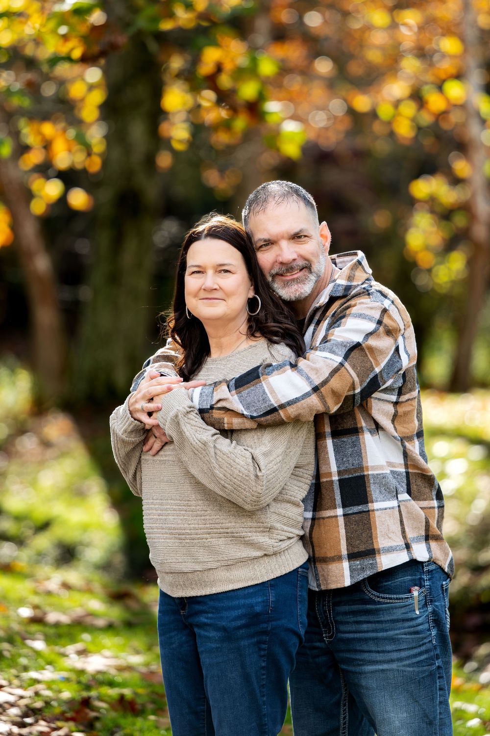 Smiling man in a plaid flannel shirt hugging a woman in a cream sweater from behind amid glowing autumn leaves.