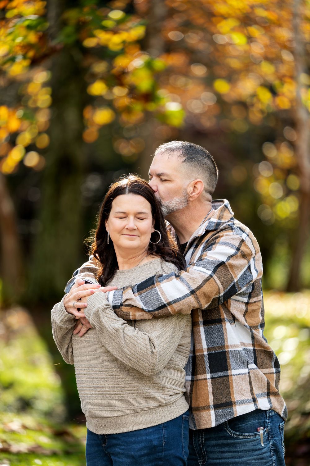 A bearded man in a plaid flannel shirt embraces and kisses a smiling woman from behind in an autumn park.