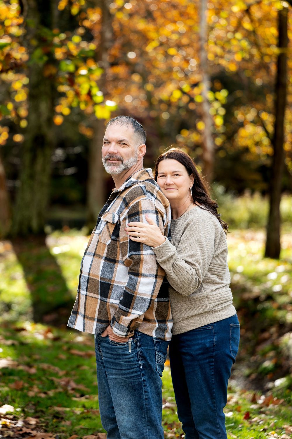 Woman in a cream sweater hugging a bearded man in a plaid shirt from behind in a golden autumn park.