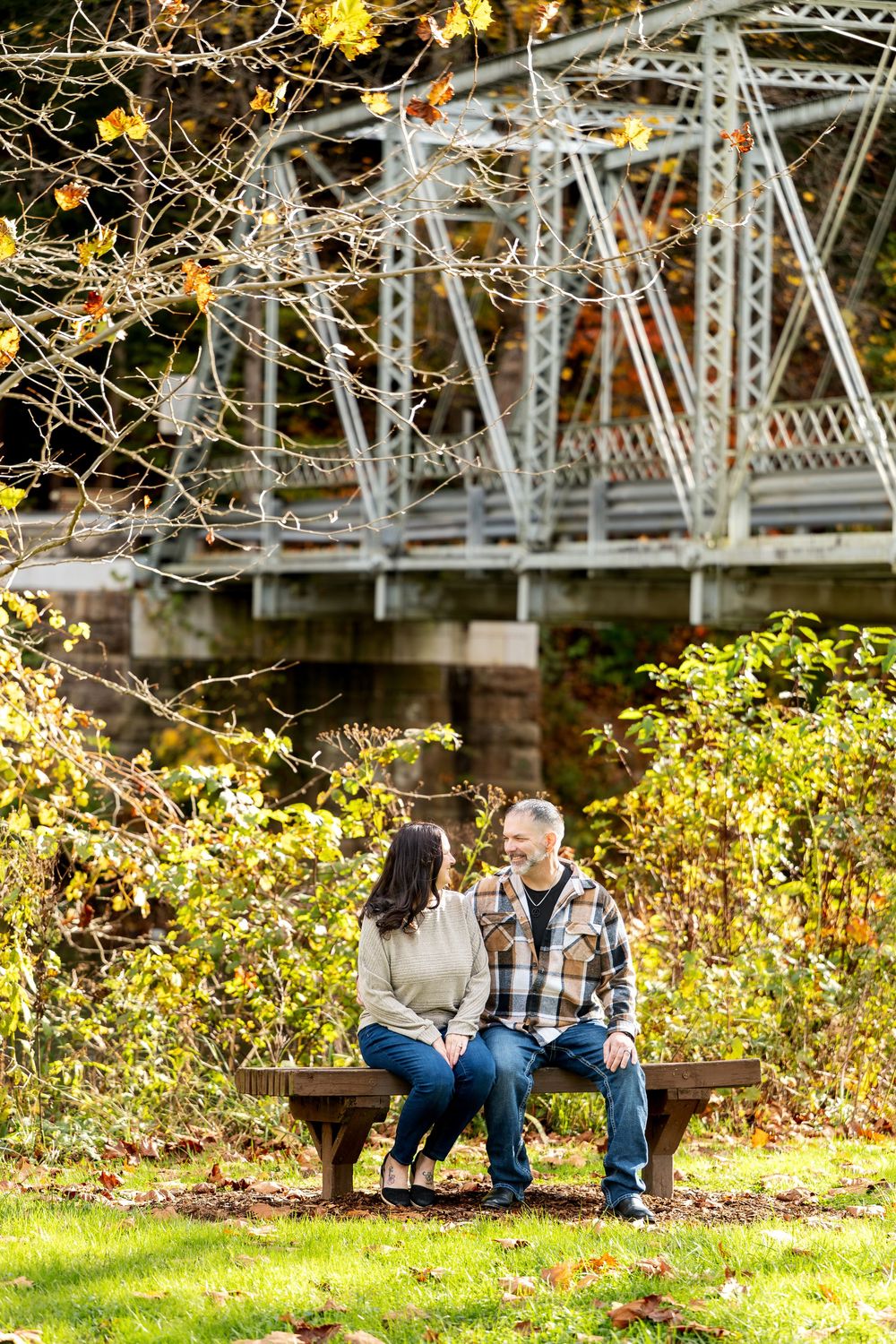 Couple sitting together on a wooden bench in a park with a steel truss bridge and autumn leaves in the background.
