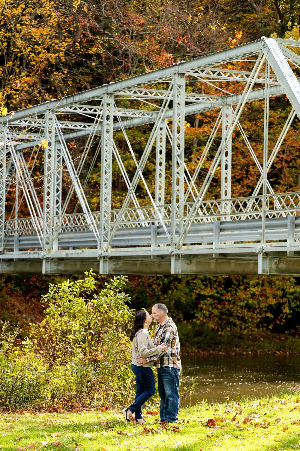Couple sharing a kiss on a grassy riverbank beneath a steel truss bridge framed by vibrant orange and yellow fall foliage.