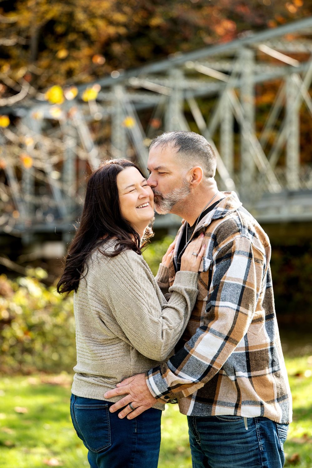 A smiling couple embraces as a man kisses a woman's cheek in an autumn park with an old steel bridge behind them.