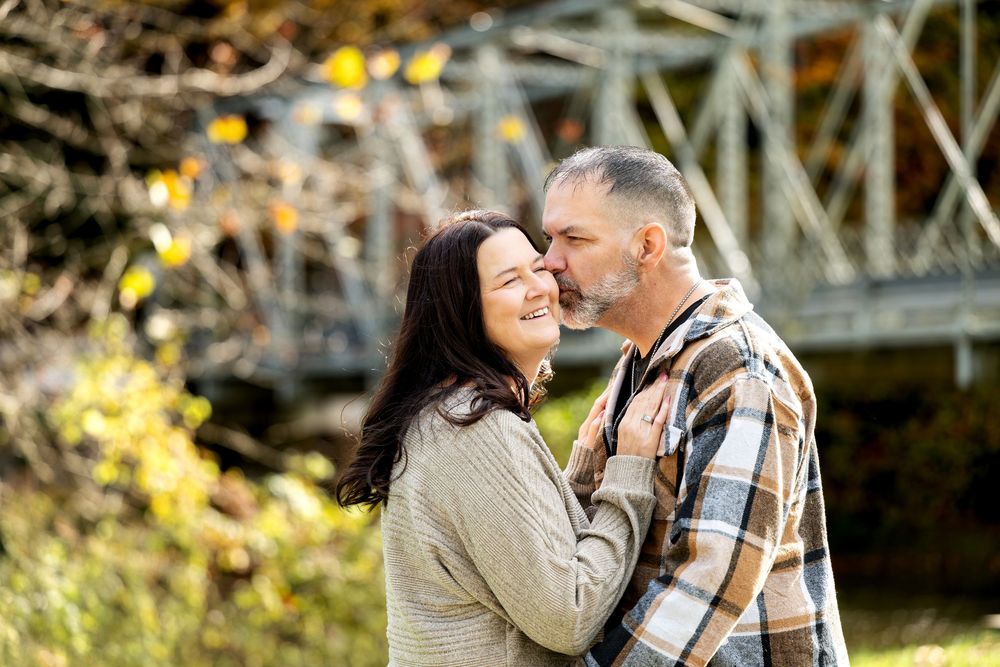 Smiling couple in casual sweater and plaid flannel share an affectionate moment outdoors near a bridge surrounded by golden autumn foliage.