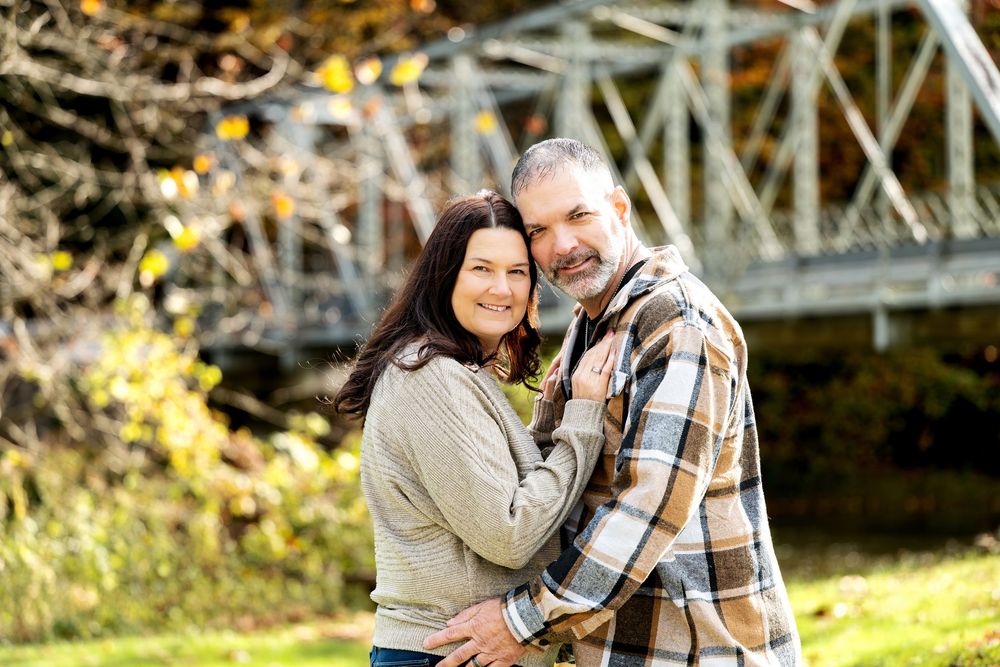 Smiling couple embracing outdoors in autumn, wearing cozy sweater and plaid flannel shirt, with a steel bridge and fall foliage behind them.