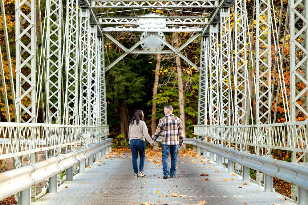 Couple holding hands walking across a historic steel truss bridge surrounded by colorful autumn foliage.