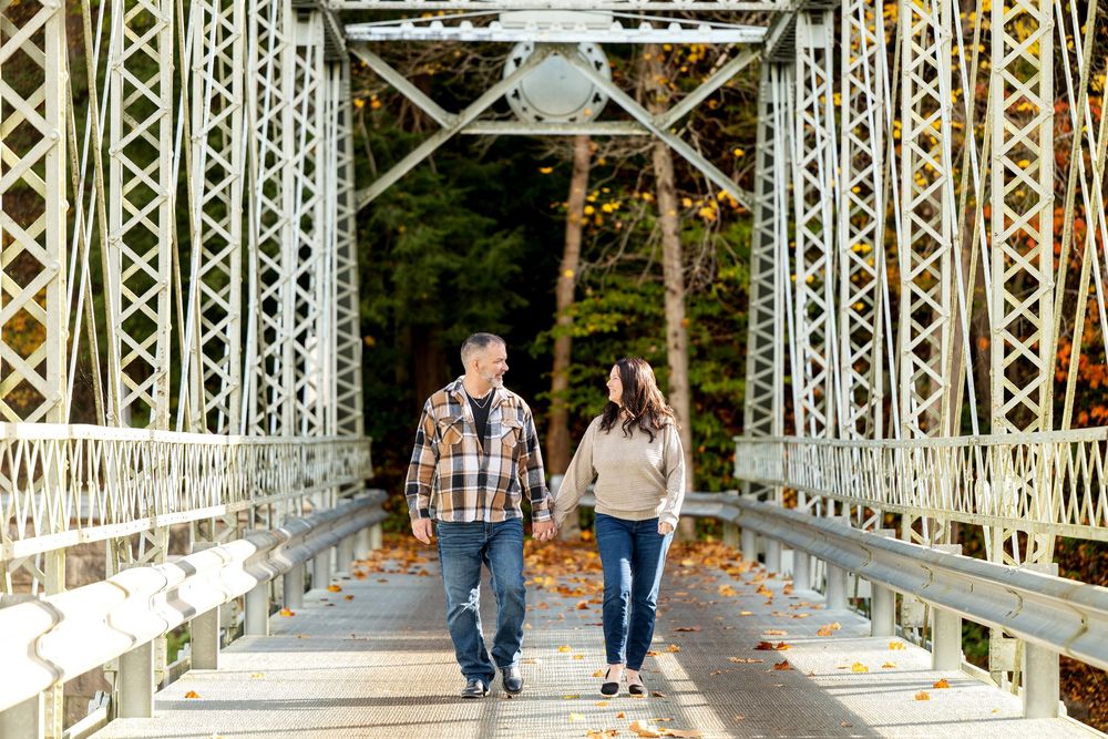 Couple in casual fall clothes holding hands and walking across a historic white steel truss bridge surrounded by autumn trees.