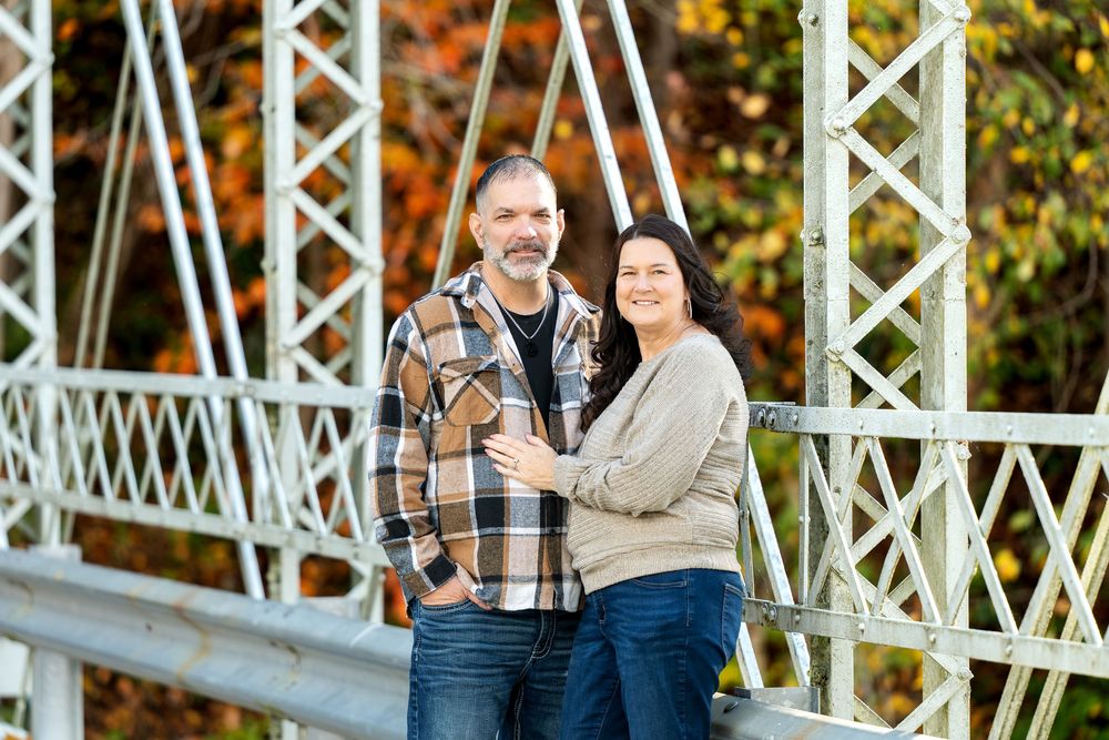 Smiling couple in casual clothes embracing on a white metal truss bridge with colorful autumn foliage in the background.