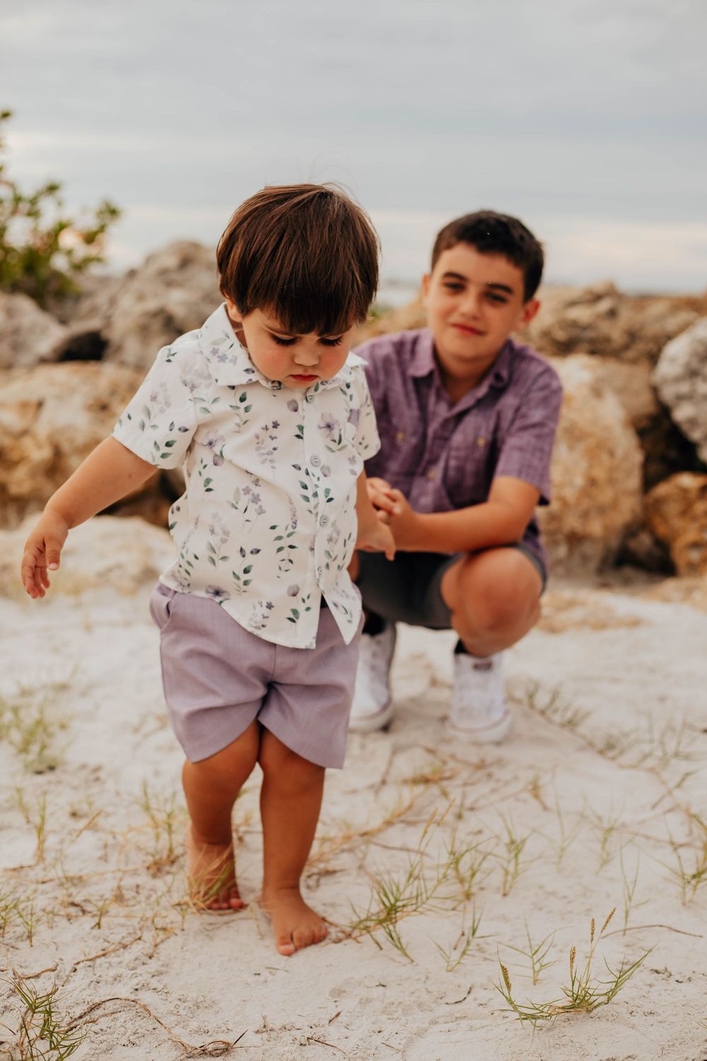 A barefoot toddler in a floral shirt and lilac shorts walks across sandy ground as an older boy crouches behind him near rocks.