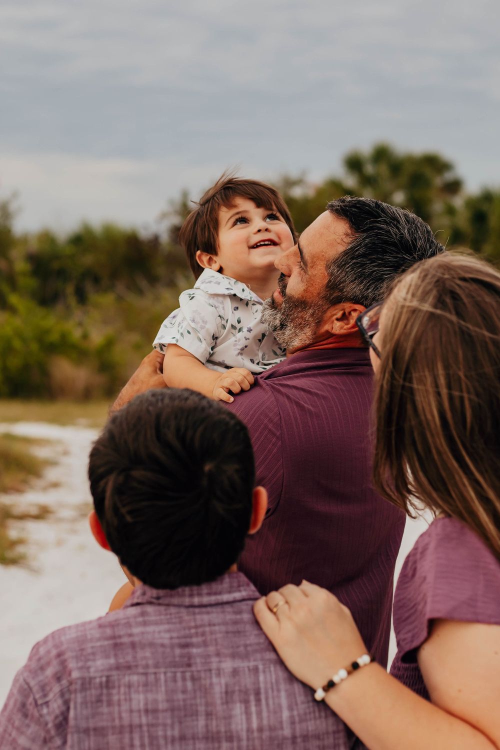 A smiling toddler is lifted in a father's arms as the family gathers outdoors on a sandy path with greenery in the background.