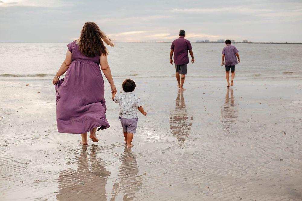 Family of four in purple outfits walking barefoot along a wet sandy beach at sunset, with a mother holding her toddler's hand.