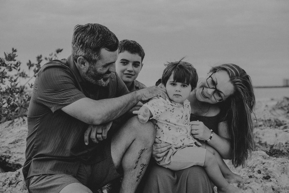 Black and white photo of a smiling family of four with a toddler on mom's lap, sitting together on a sandy beach.