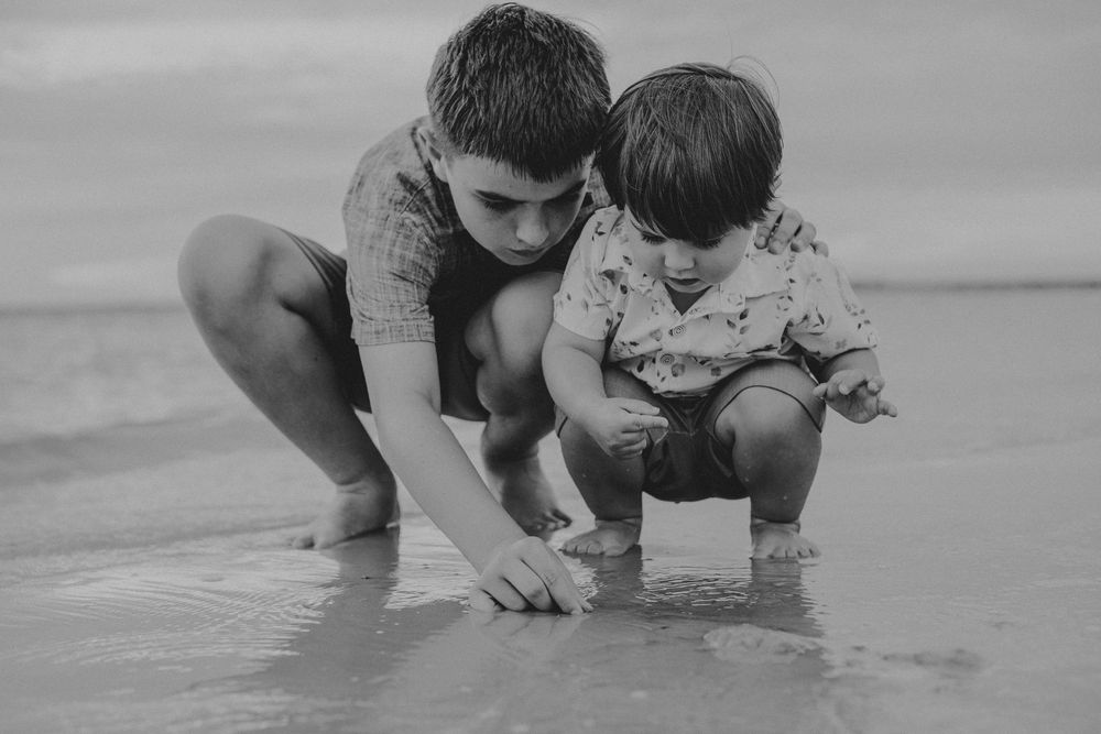 Two young children crouch together at the water's edge on a beach, curiously touching the wet sand in this black and white photo.