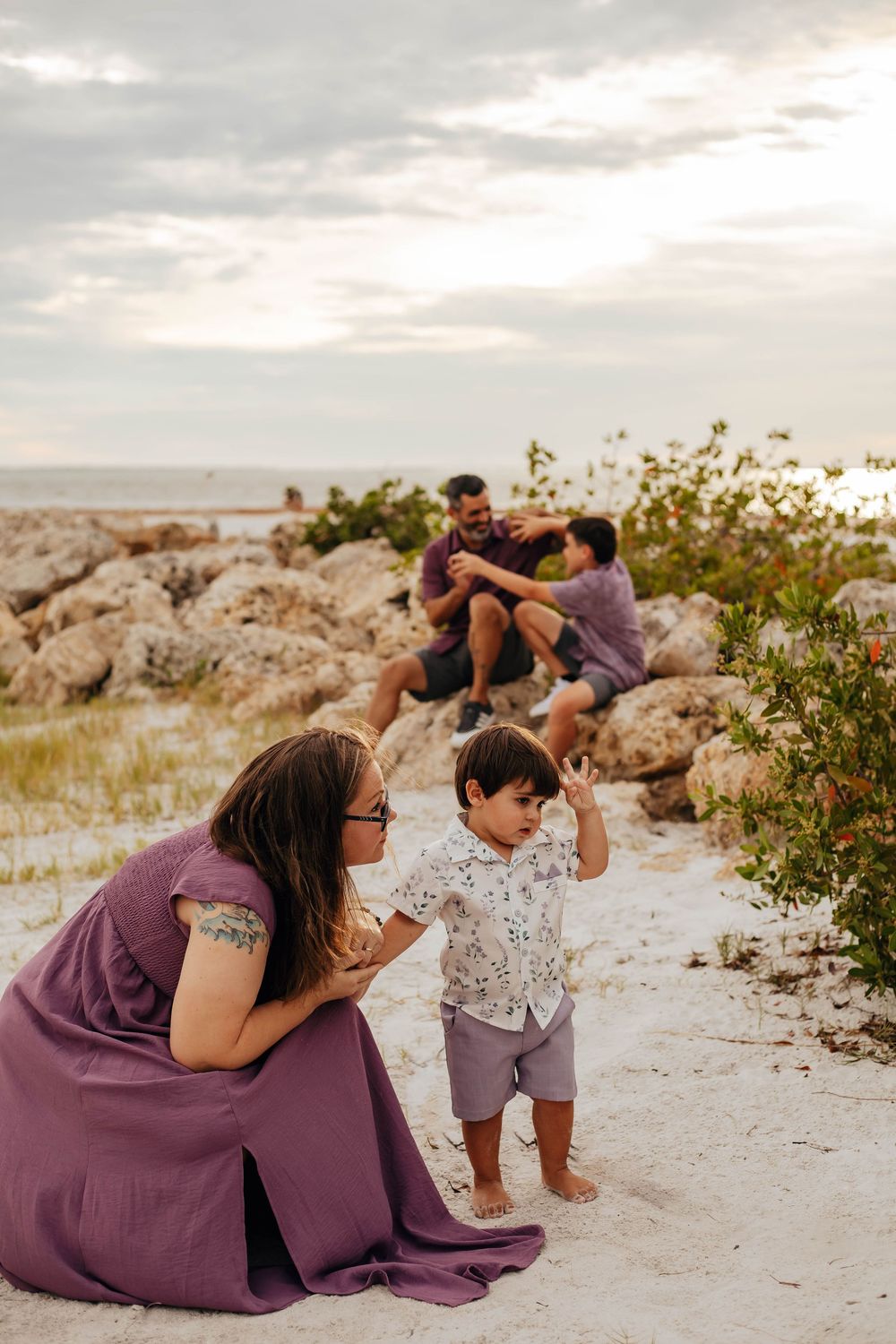 Family beach photo at sunset with a woman in a purple dress holding a toddler's hand while a man and boy sit on rocks behind them.