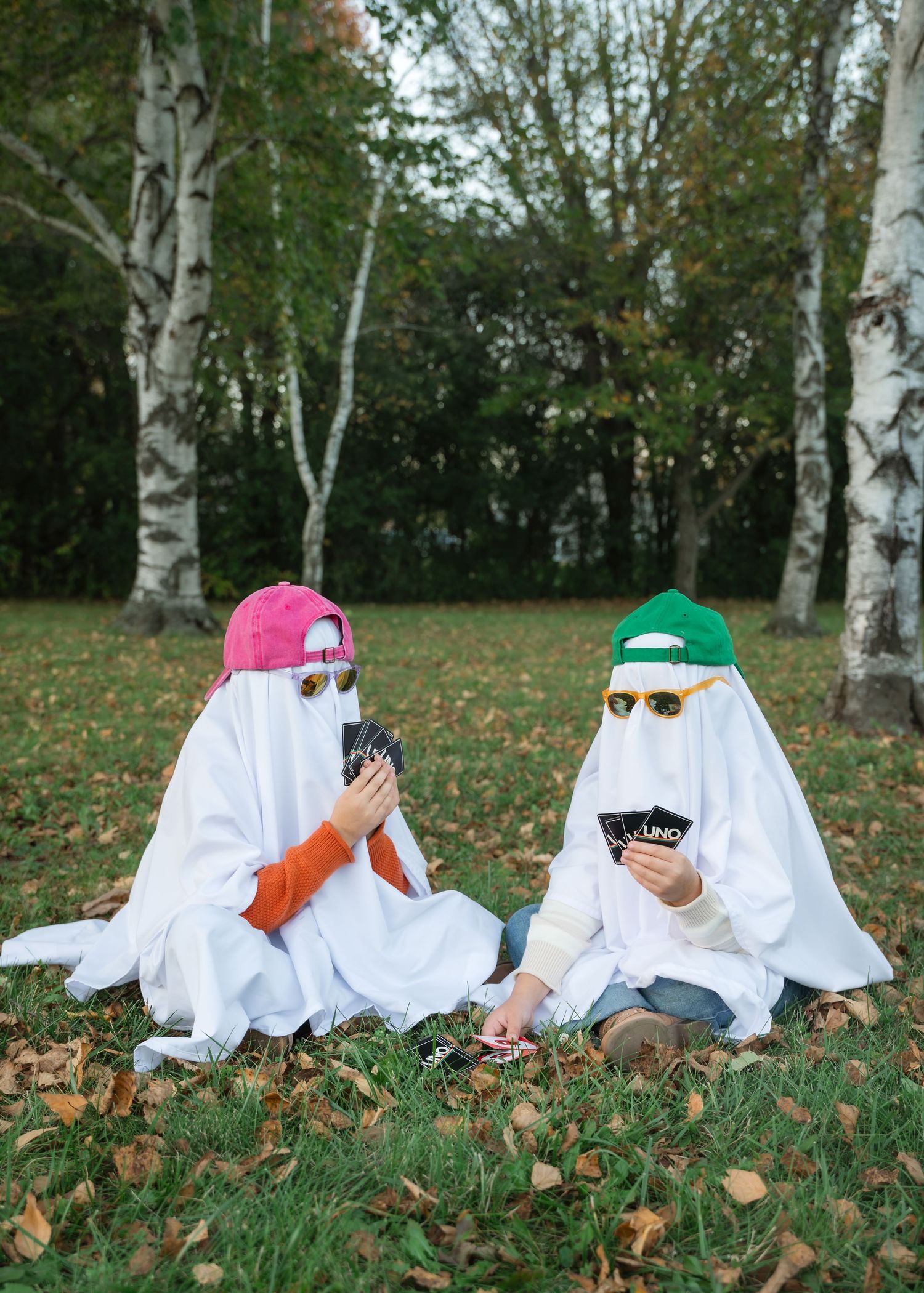 Two people in white ghost sheets with baseball caps and sunglasses sit on autumn grass playing Uno cards beneath birch trees.