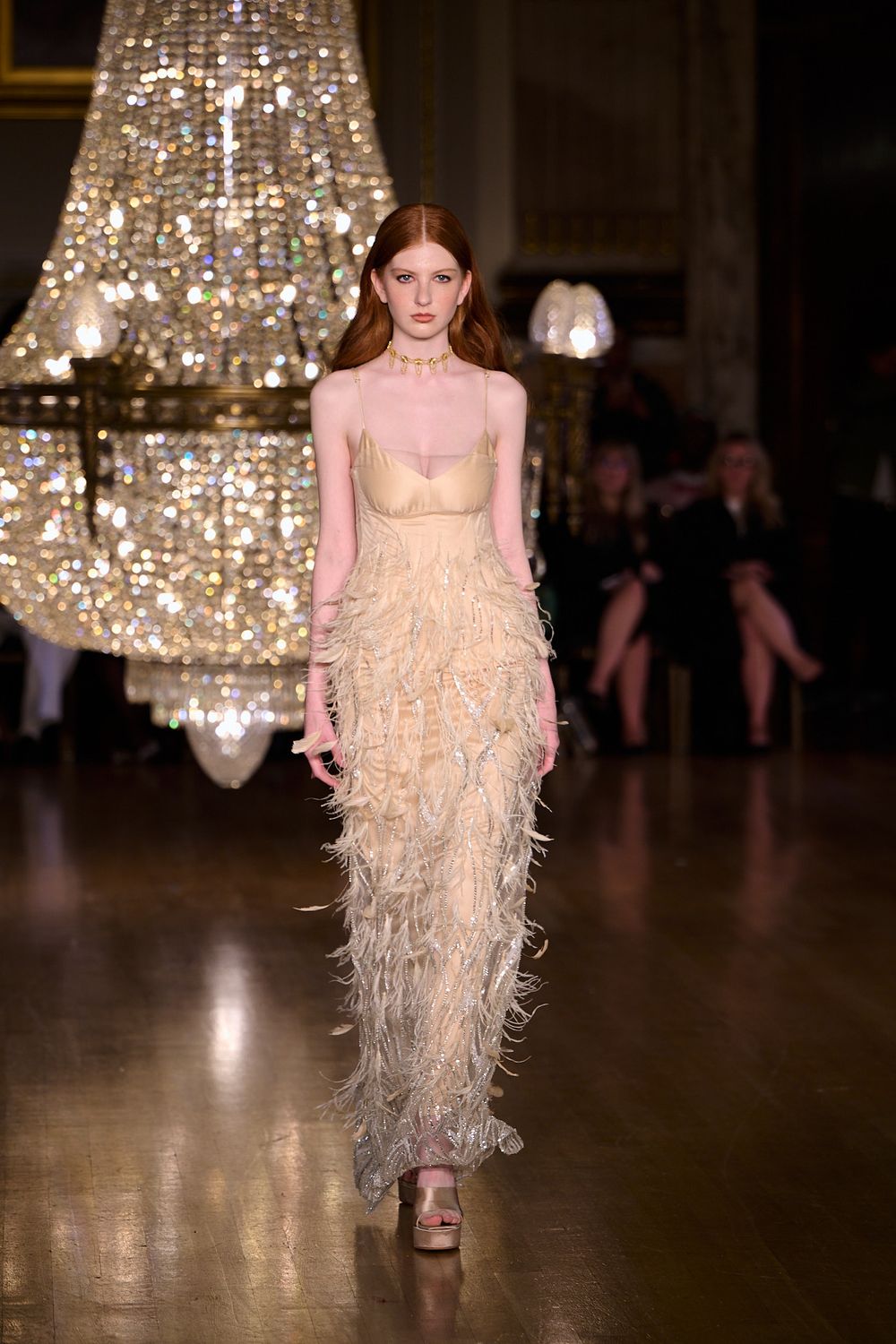Model walks a runway in a champagne feathered evening gown with spaghetti straps, beneath a sparkling crystal chandelier, as guests watch.