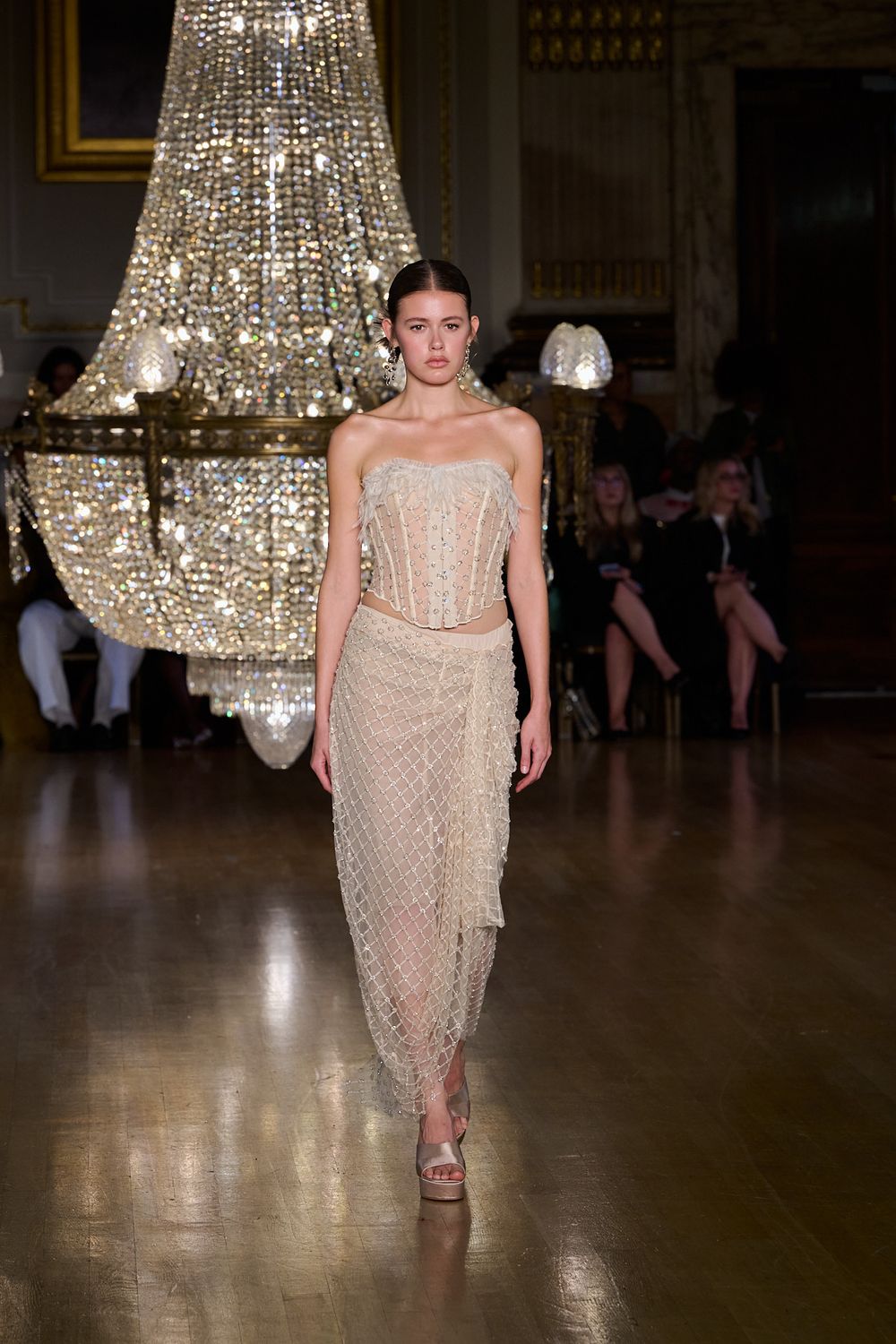 Model walks a runway in a beaded corset top and sheer embellished wrap skirt beneath a large crystal chandelier as guests watch.