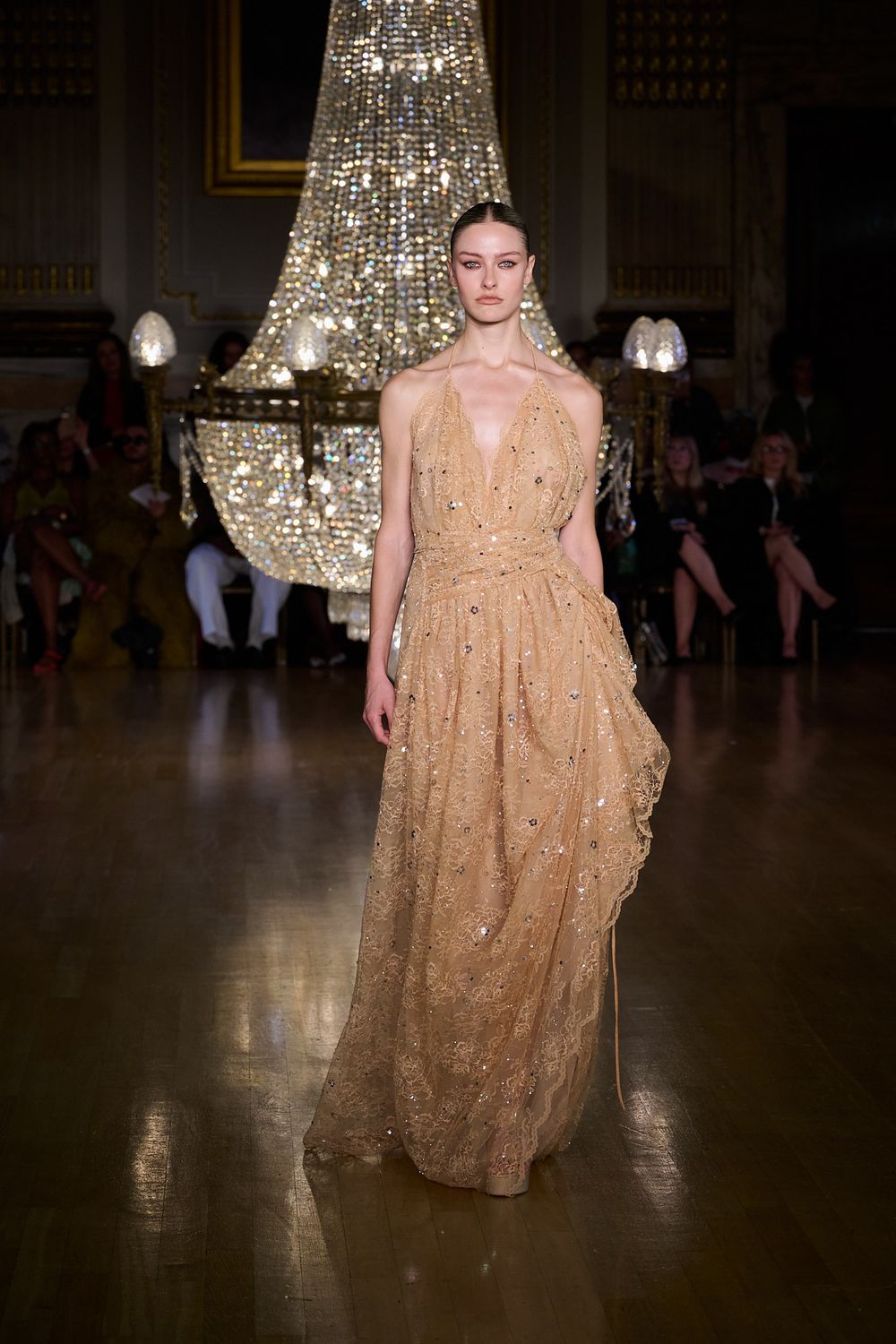 Model walks a runway in a sparkling gold sequined plunging-neckline gown beneath a grand crystal chandelier as guests watch.