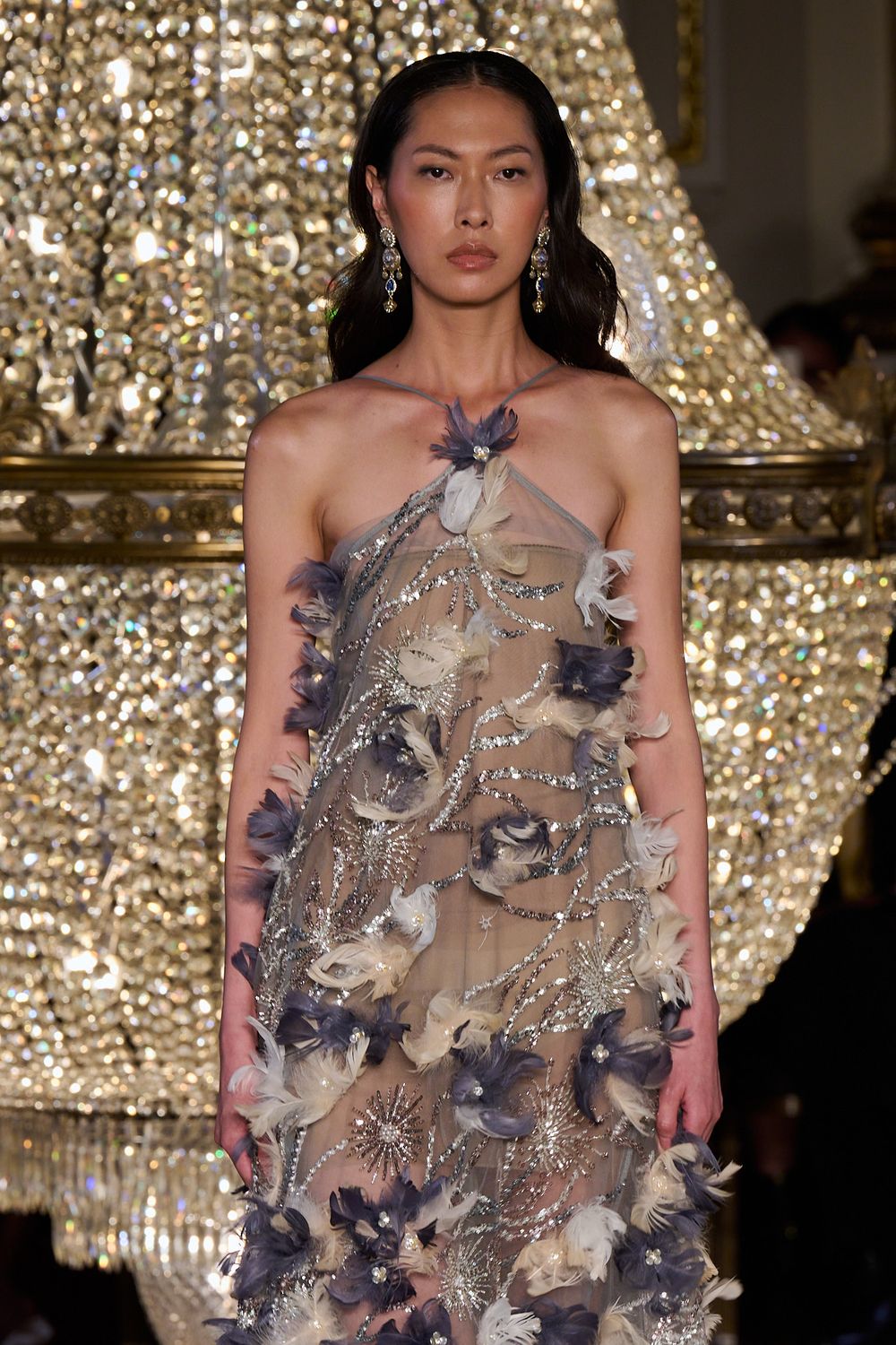 Model on the runway wears a sheer halter gown embellished with gray and cream floral appliqués, beside a glittering crystal chandelier.
