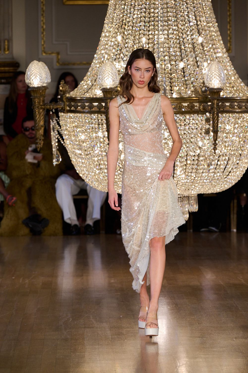 Model walks a runway in a sparkling champagne sequin dress and platform sandals beneath a grand crystal chandelier as guests watch.