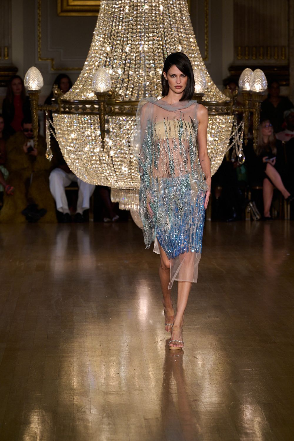 Model on the runway wears a sheer blue sequined and beaded mini dress with a yellow bodice under a sparkling crystal chandelier.