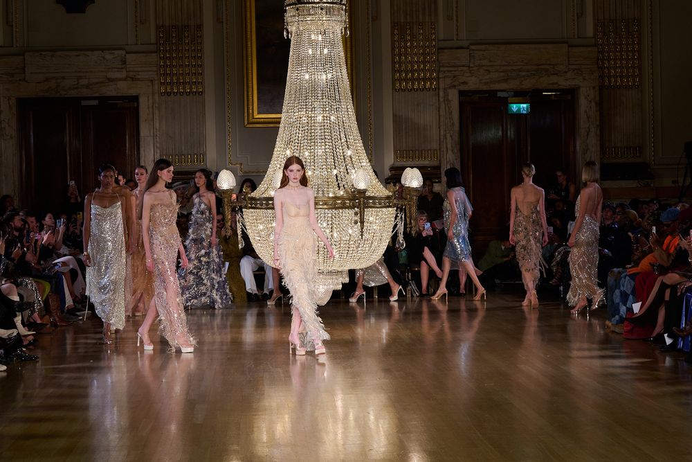 Models in flowing sheer pastel gowns walk a polished wood runway beneath a grand crystal chandelier as guests watch from the sides.