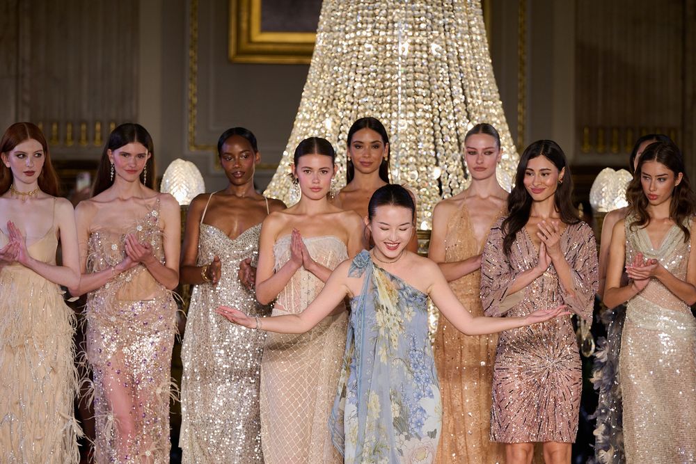 Runway models in sparkling beaded and feathered evening gowns pose together beneath a crystal chandelier at a fashion show finale.