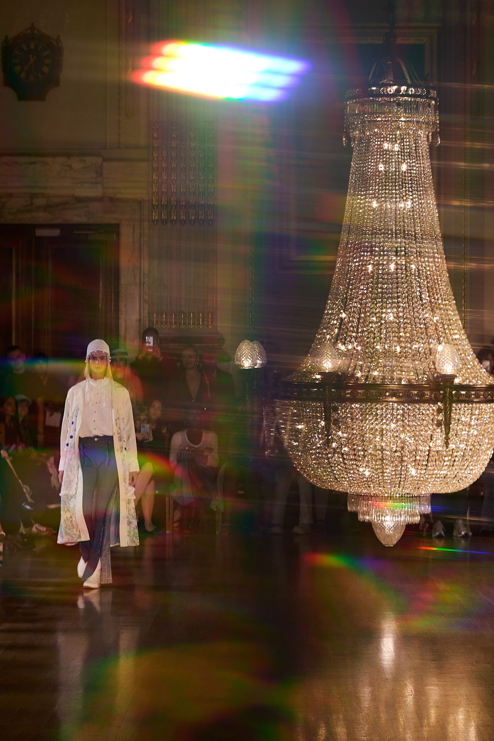 Model in a white hooded coat and dark skirt walks a runway beneath a large sparkling crystal chandelier as colorful lens flares glow.