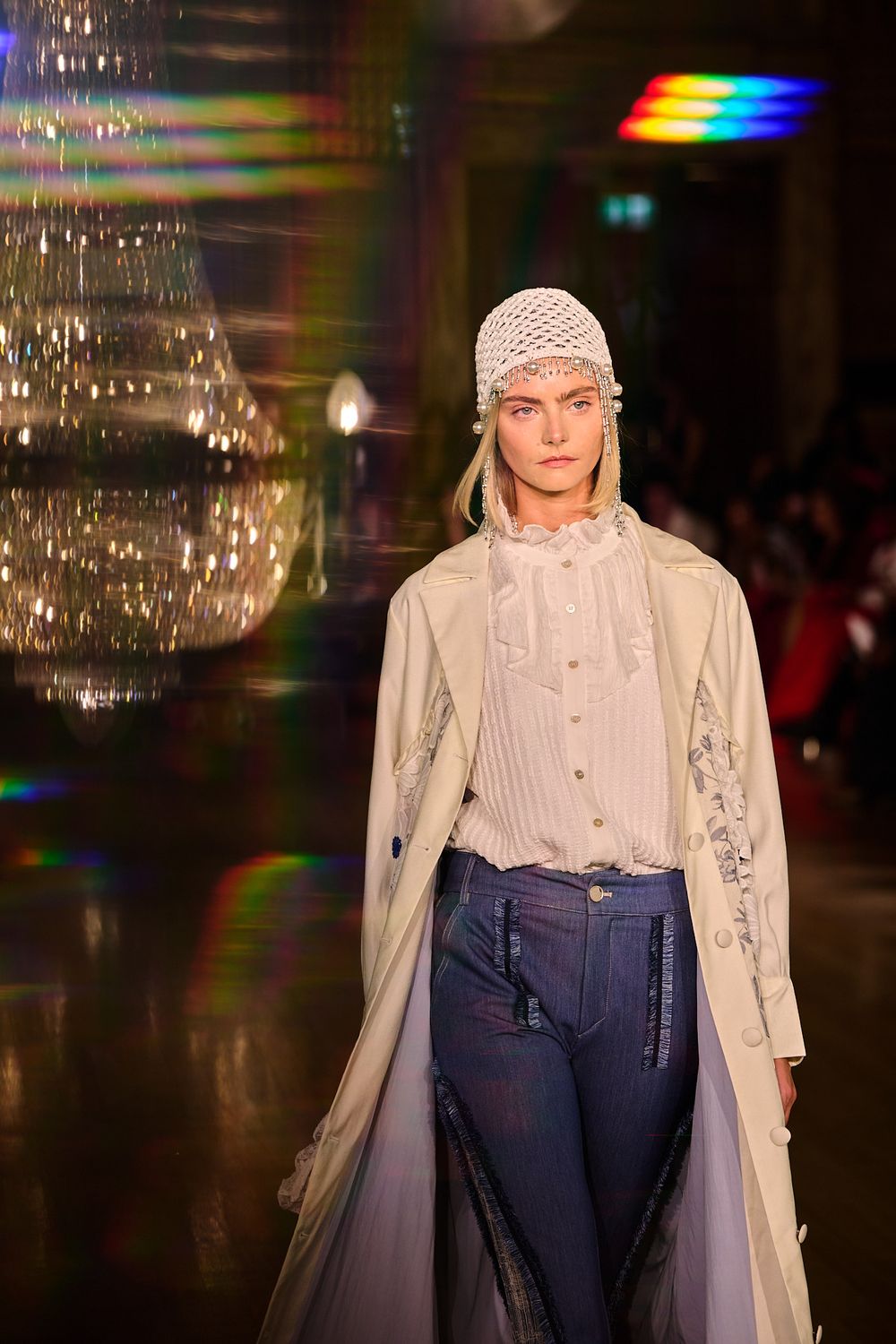 Model wearing a white crochet cap, ruffled cream blouse, long beige coat and dark jeans walks the runway near a sparkling chandelier.