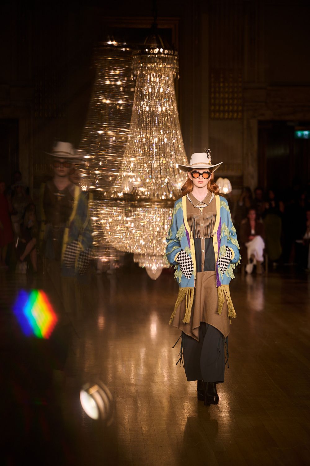 Runway model in a white cowboy hat, round glasses and a fringed blue-yellow jacket walks beneath glittering crystal chandeliers.