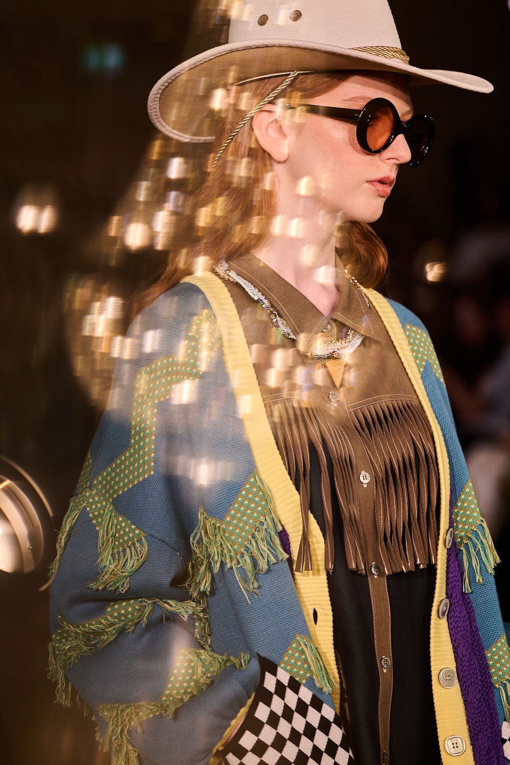 Model on a runway wears a cream wide-brim hat, round tinted sunglasses and a fringed western-style cardigan with geometric patterns.