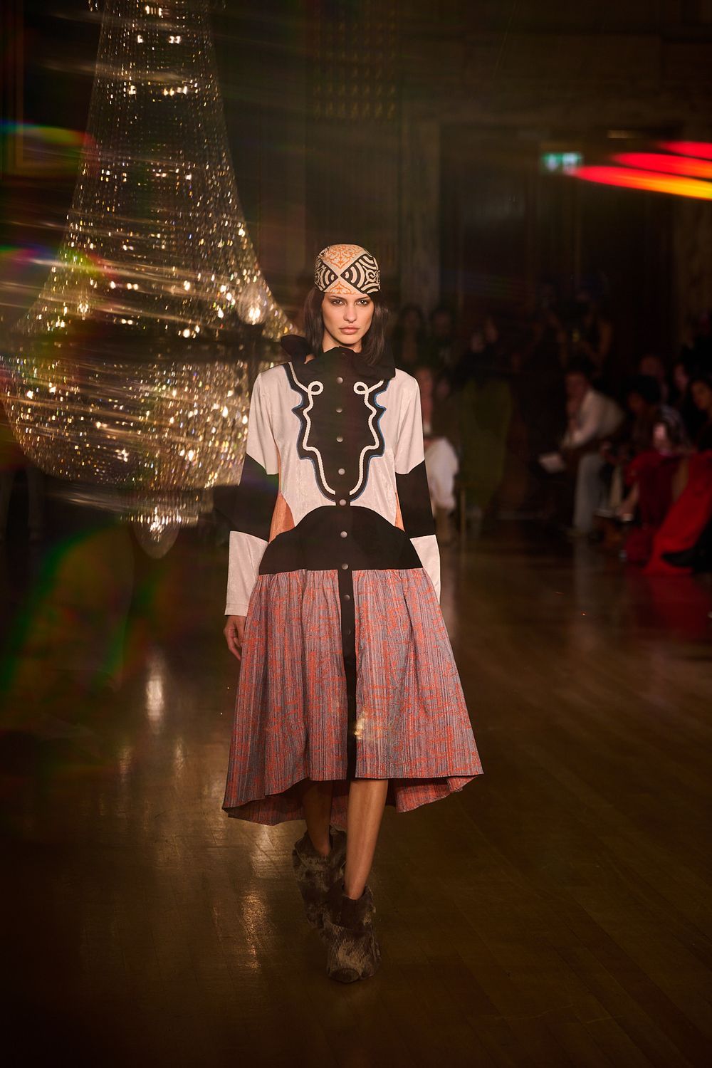 Model walks the runway in a patterned headwrap, black and cream embroidered jacket and pink plaid skirt beside a sparkling chandelier.