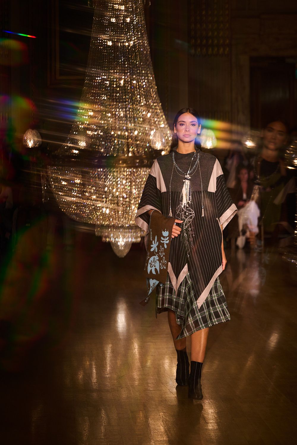Model walks a runway in an ornate hall wearing a layered black poncho, plaid skirt and black boots beneath a glittering crystal chandelier.