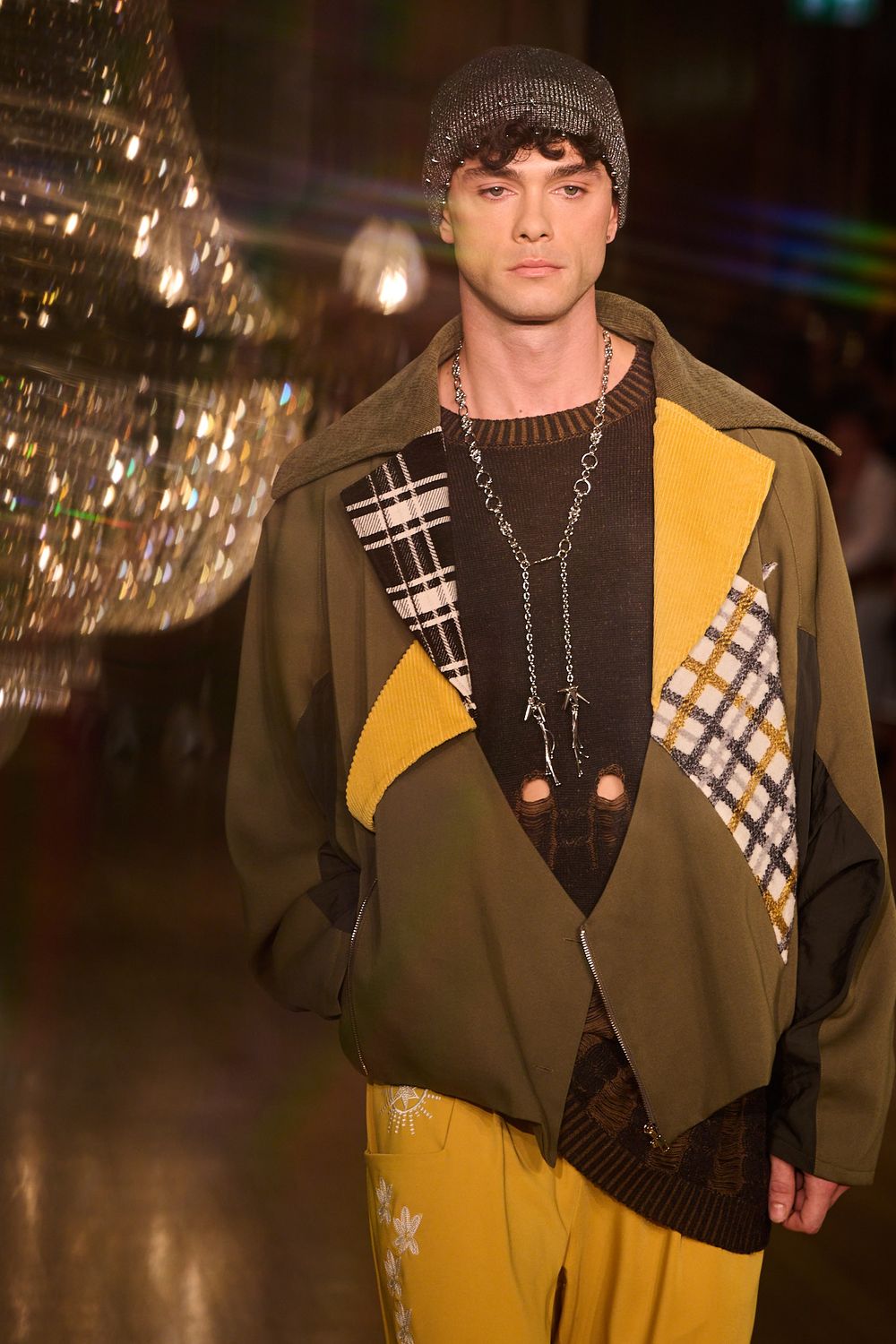 Runway model wears an olive green patchwork jacket with yellow and plaid panels, a knit beanie, chain necklace and mustard trousers.