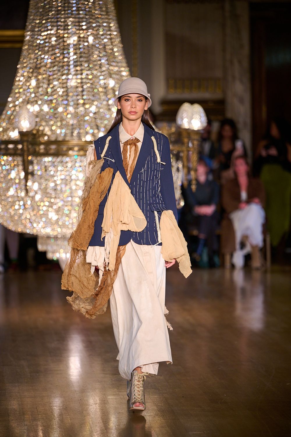 Runway model wears a gray bucket hat, navy pinstripe jacket, layered tan scarves and wide-leg cream trousers beneath a crystal chandelier.