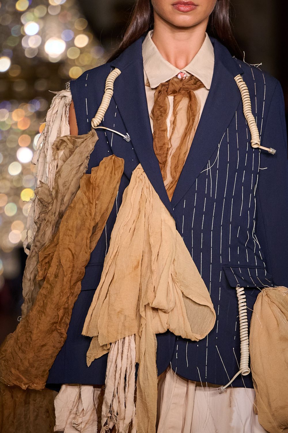 Runway model wearing a navy pinstripe blazer with draped tan silk scarves, rope toggle details and a knotted brown neck scarf.