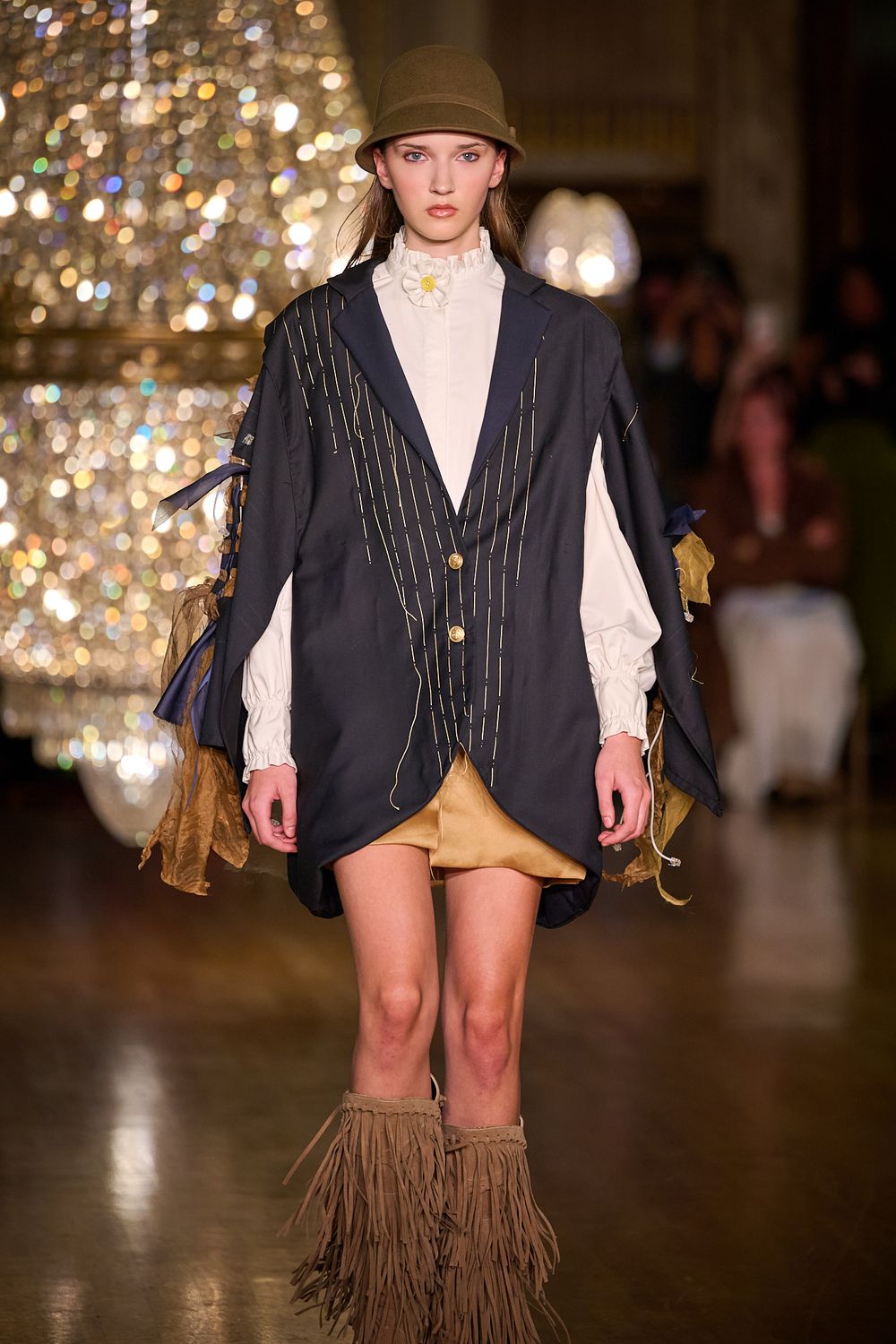 Runway model wears a brown bucket hat, navy pinstripe jacket over a white ruffled blouse, gold mini skirt and fringed suede boots.