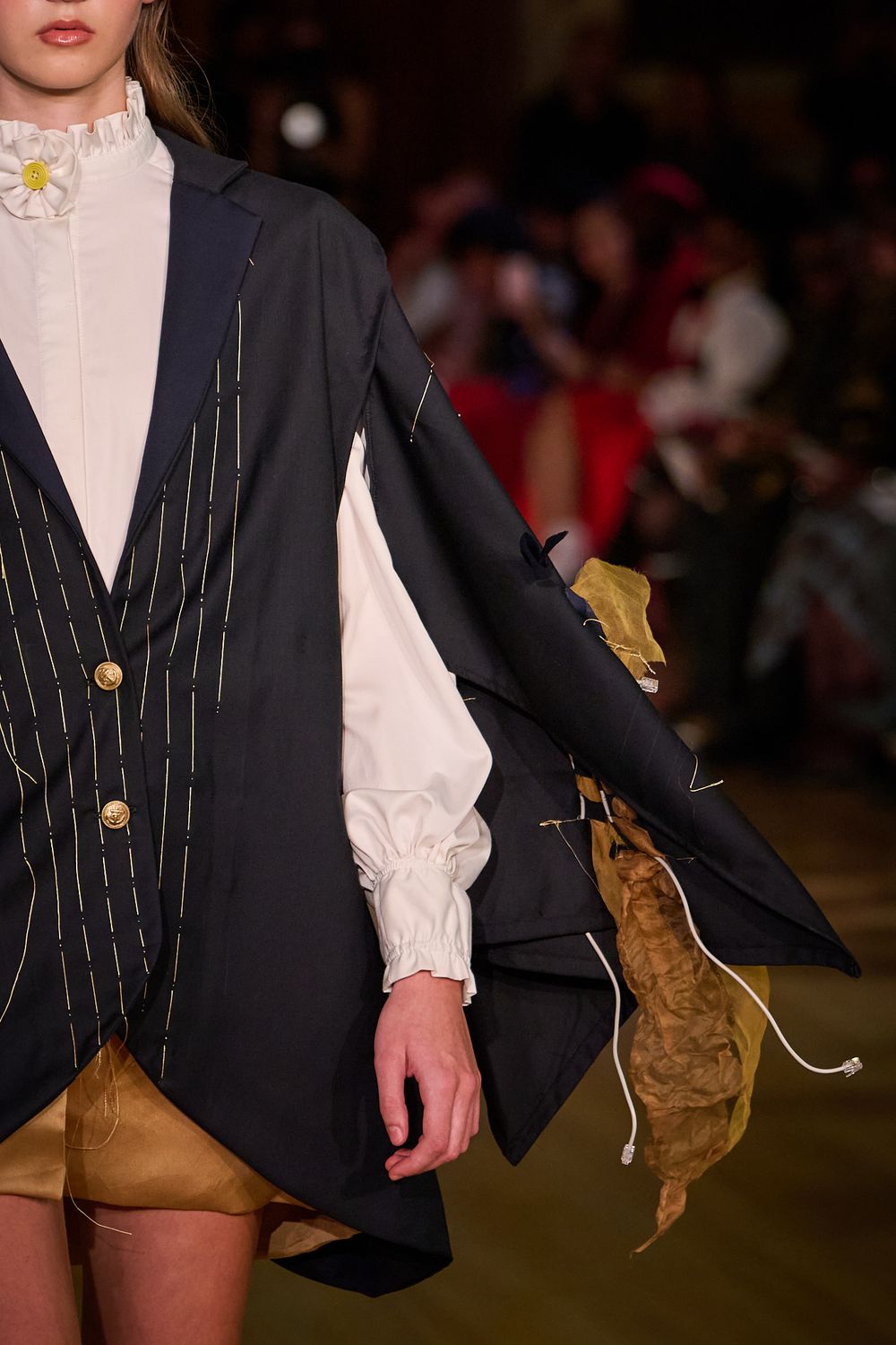 Runway model wears a navy pinstripe asymmetric jacket over a white ruffled blouse with a floral collar pin and gold shorts.