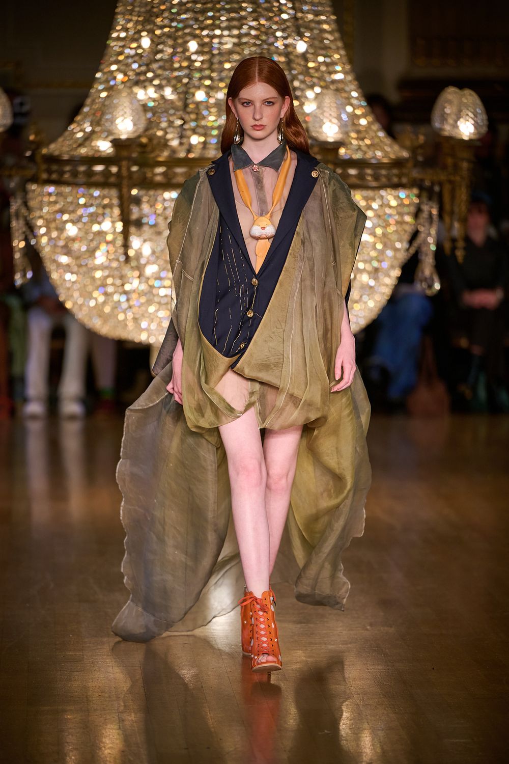 Red-haired model walks the runway in an olive sheer layered coat, navy vest, shorts and orange boots beneath a sparkling crystal chandelier.