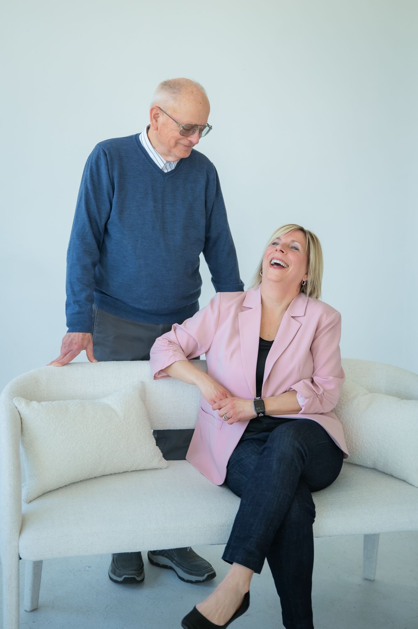 An older man in a blue sweater stands behind a woman in a pink blazer sitting on a white sofa, both laughing.