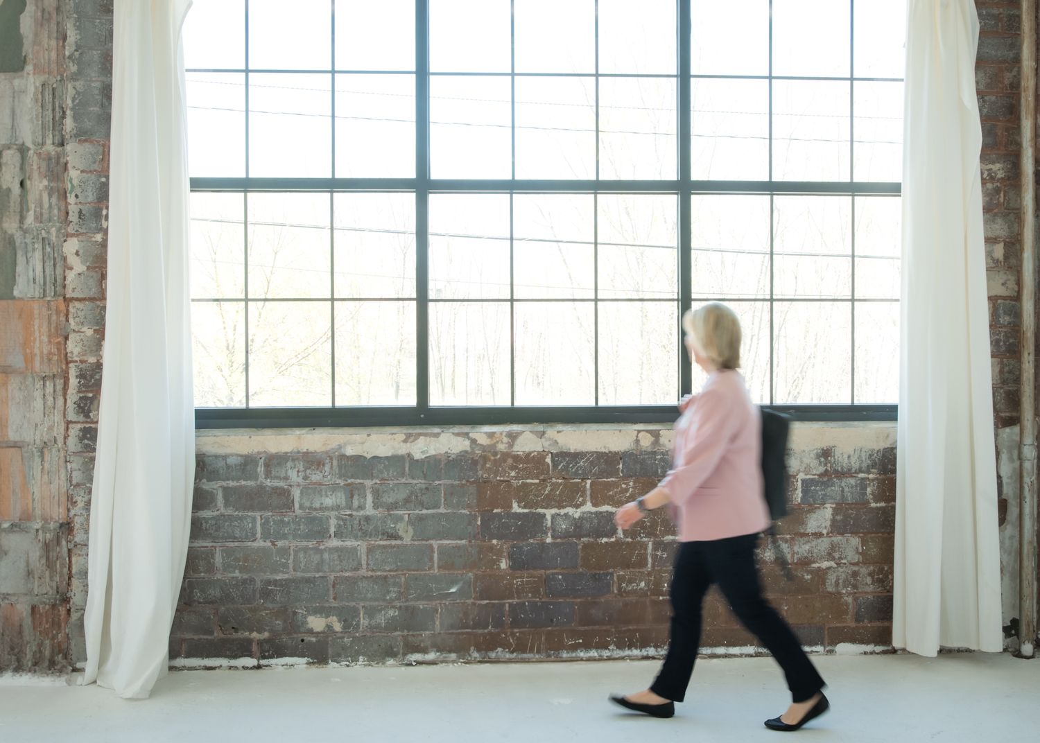 A woman in a pink sweater and black pants walks past large industrial windows in a bright brick-walled studio space.