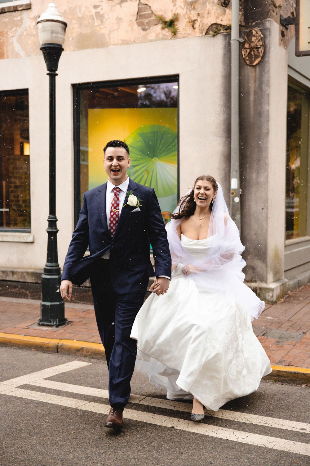 Joyful newlyweds hold hands crossing a street, bride in white gown with veil, groom in navy suit with red tie.