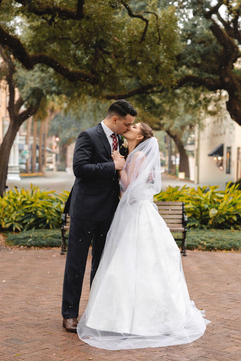 Bride and groom share a romantic kiss outdoors in a park, bride in white gown with veil, groom in dark suit.