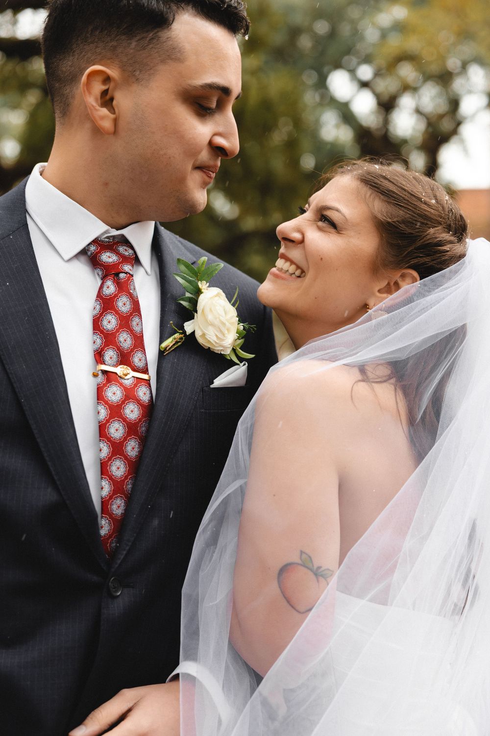 Smiling bride in veil and groom in gray suit with red tie share a romantic moment outdoors on their wedding day.