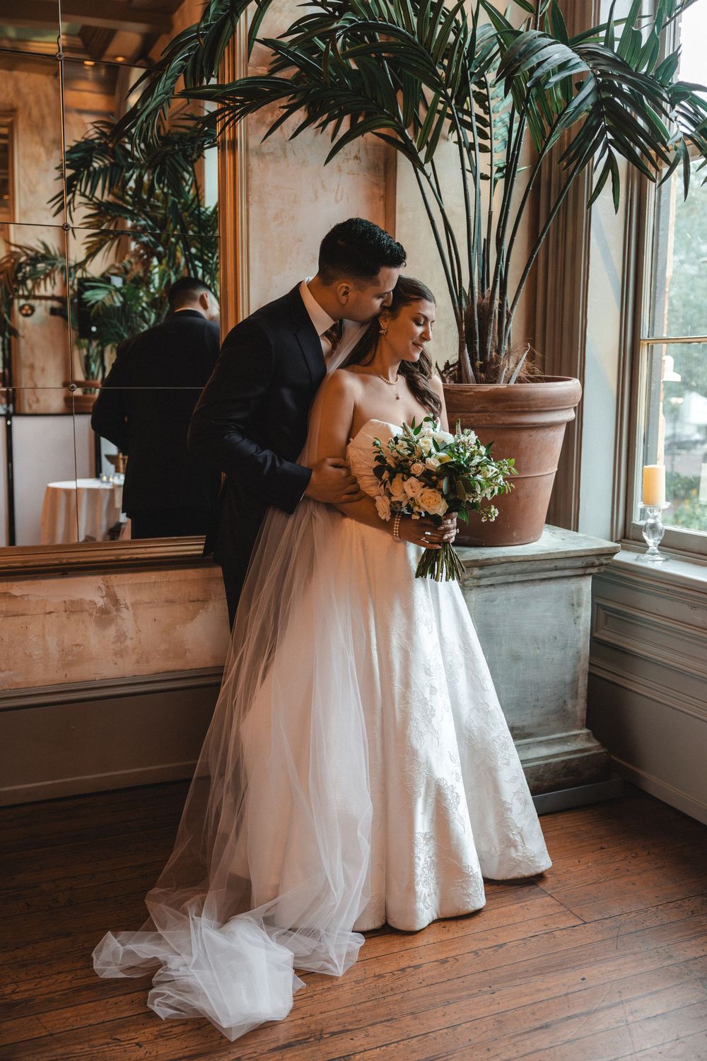 Bride in lace gown holding bouquet stands with groom in dark suit, sharing a tender moment by window with tropical plants.