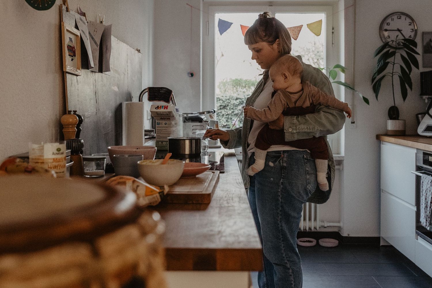 Parent multitasking in kitchen while carrying baby in carrier, preparing food near wooden countertop with natural light.