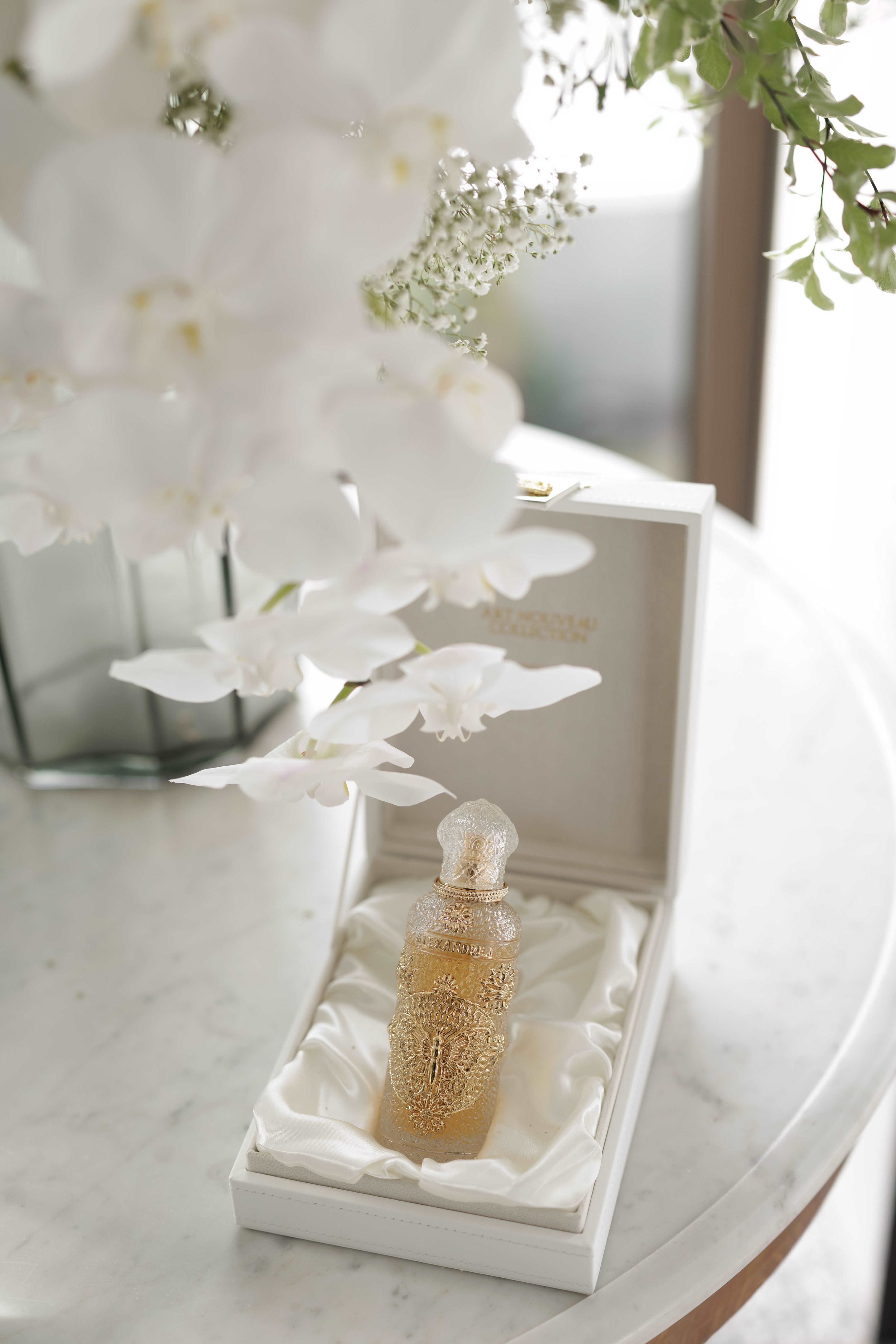Luxury perfume bottle in ornate gold filigree displayed in white satin box beside white orchids on marble table.