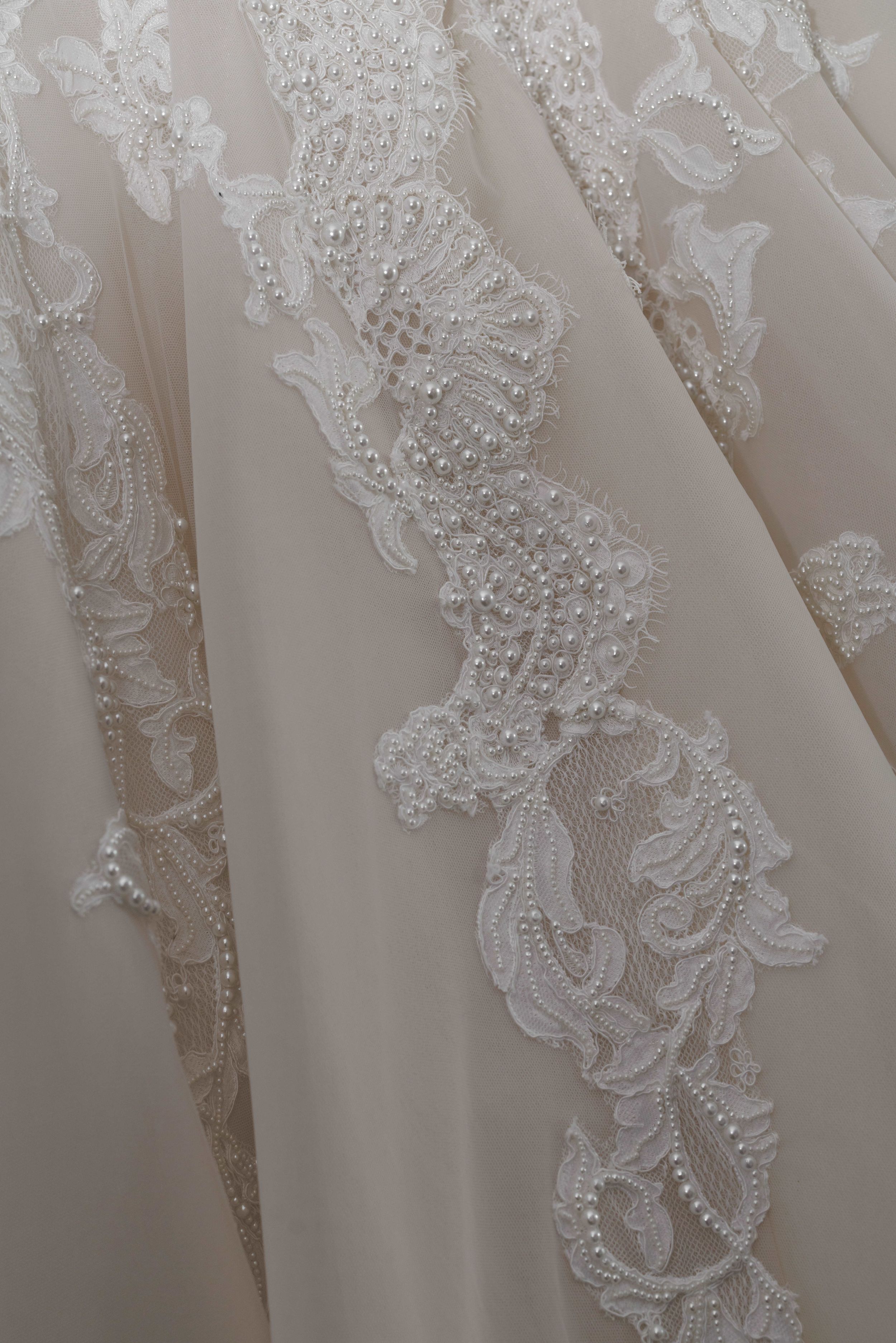 Close-up of elegant white wedding dress featuring intricate lace appliqué with pearl beading on flowing chiffon fabric.